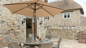 A stone building with a thatched roof, a round table with wicker chairs, and a large umbrella. - The Farmhouse at Higher Westwater (Axminster)