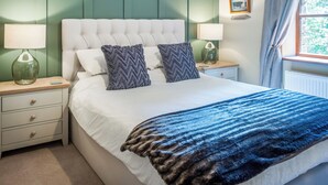 A bedroom with a tufted headboard, two bedside tables with lamps, a bed with a blue and brown striped blanket, and a window with a curtain. - An attractive and spacious cottage close to the River Eamont. (Pooley Bridge, nr. Ullswater)