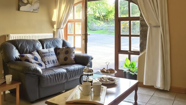 A cozy living room with a leather sofa, a coffee table with a tray of pastries, and a view of a green landscape through a large window. - 2 bedroom accommodation in Glynarthen, near Cardigan (Glynarthen, near Cardigan)