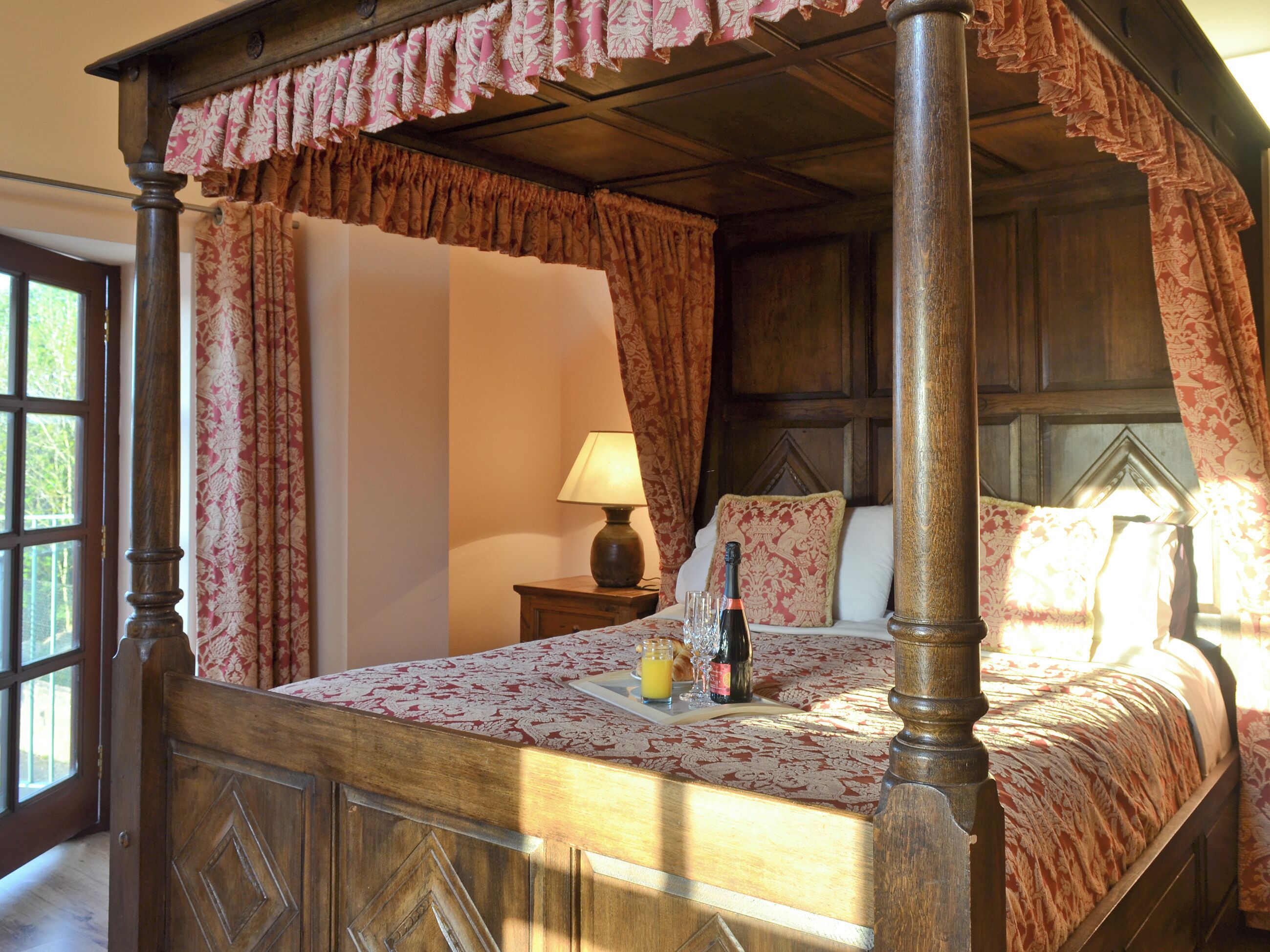 A four-poster bed with a floral bedspread, a bedside table with a lamp, and a bottle of champagne.