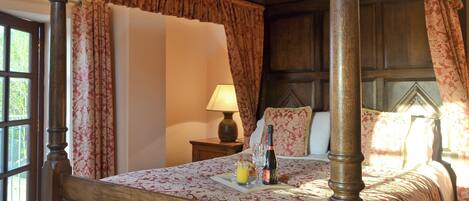 A four-poster bed with a floral bedspread, a bedside table with a lamp, and a bottle of champagne.