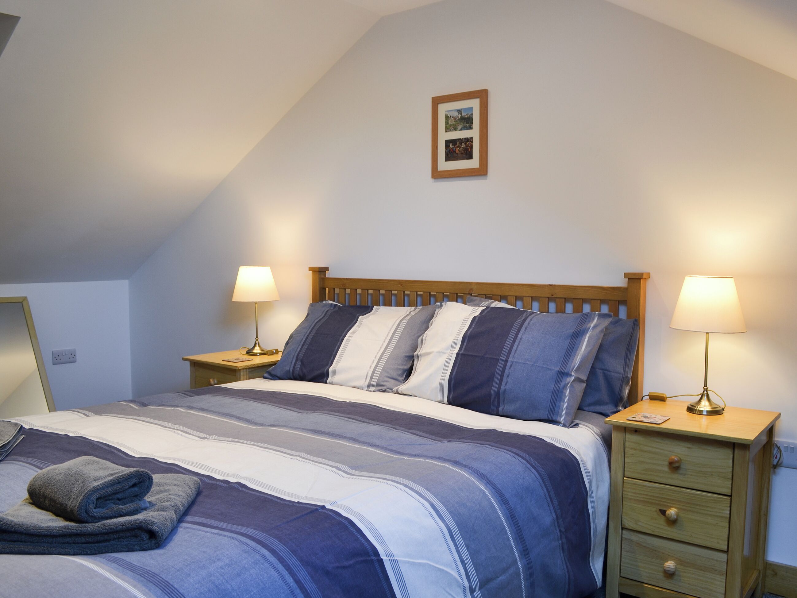 A neatly made bed with striped bedding, two bedside lamps, a wooden nightstand, and a framed picture on the wall.
