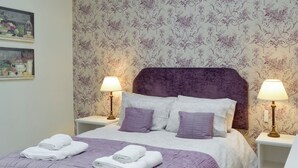 A neatly made bed with a purple bedspread and pillows, two bedside lamps, and a floral wallpaper pattern. - 4 bedroom accommodation in East Allington, near Totnes (East Allington, near Totnes)