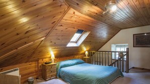 Un dormitorio con techo inclinado, un tragaluz, una cama con colcha verde, dos lámparas de noche, un aparador de madera, una silla de mimbre y un cubo de basura pequeño.