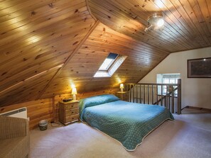 Une chambre avec un plafond en pente, un puits de lumière, un lit recouvert d’une couverture verte, deux lampes de chevet, une commode en bois, une chaise en osier et une petite poubelle.