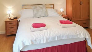 A neatly made bed with white bedding, two pillows, and two pink towels. There are two bedside tables with lamps, a wooden wardrobe, and a wooden nightstand with drawers. - Russet Cottage - E4678 (Near Lympsham)