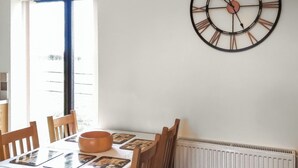 A dining area with a table set for two, a wall clock, and a window with blinds. - Russet Cottage - E4678 (Near Lympsham)