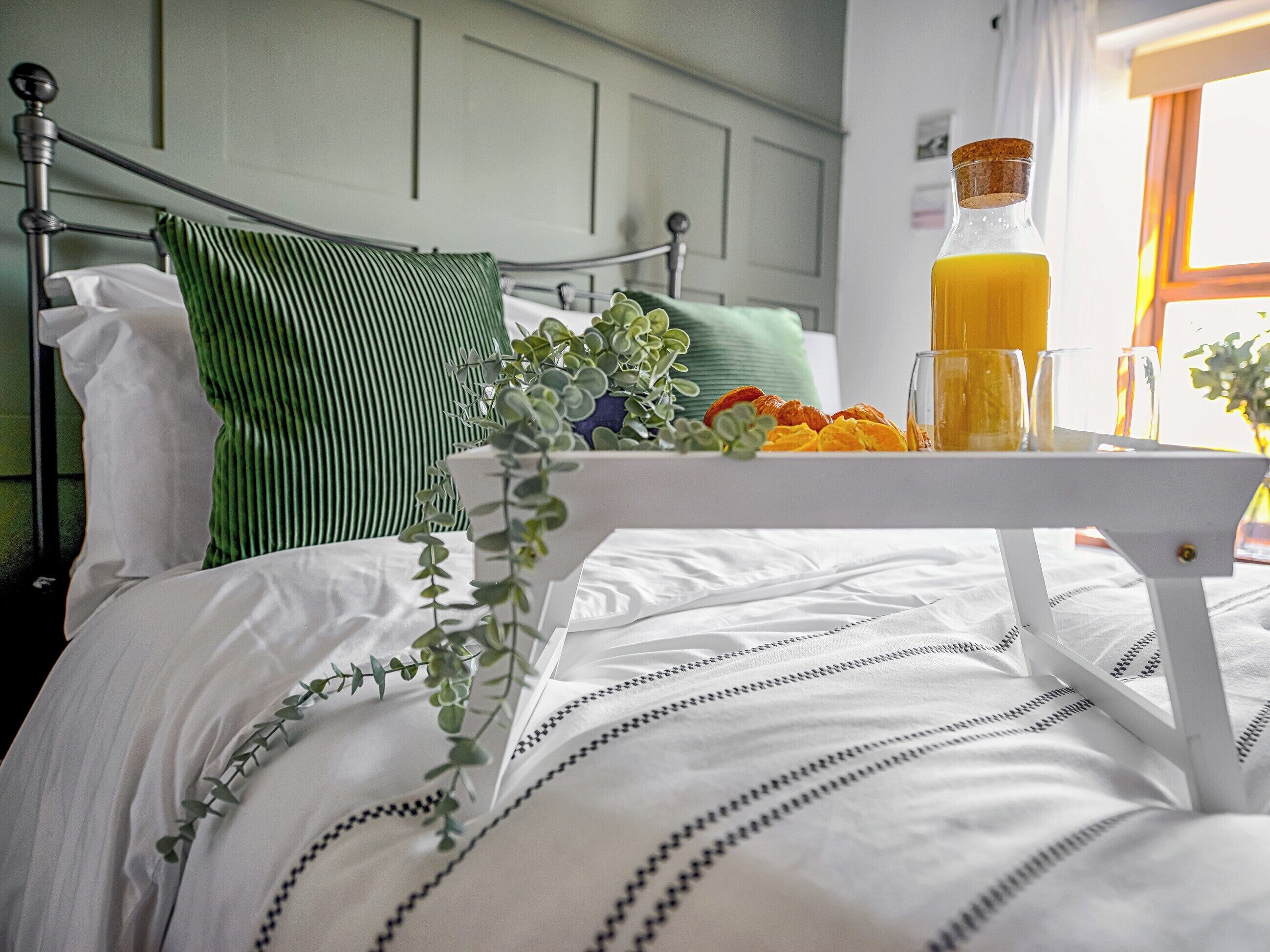 A neatly made bed with a white and black striped bedspread, a green pillow, and a white bedside table with a glass of orange juice and a bottle.