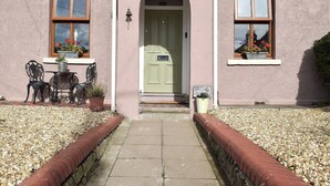 Ein Gebäude mit einer grünen Tür, zwei Fenstern mit Blumenkästen, einem Schild mit der Aufschrift "Moss Cottage" und einem gepflasterten Eingang mit roter Ziegelbordüre.