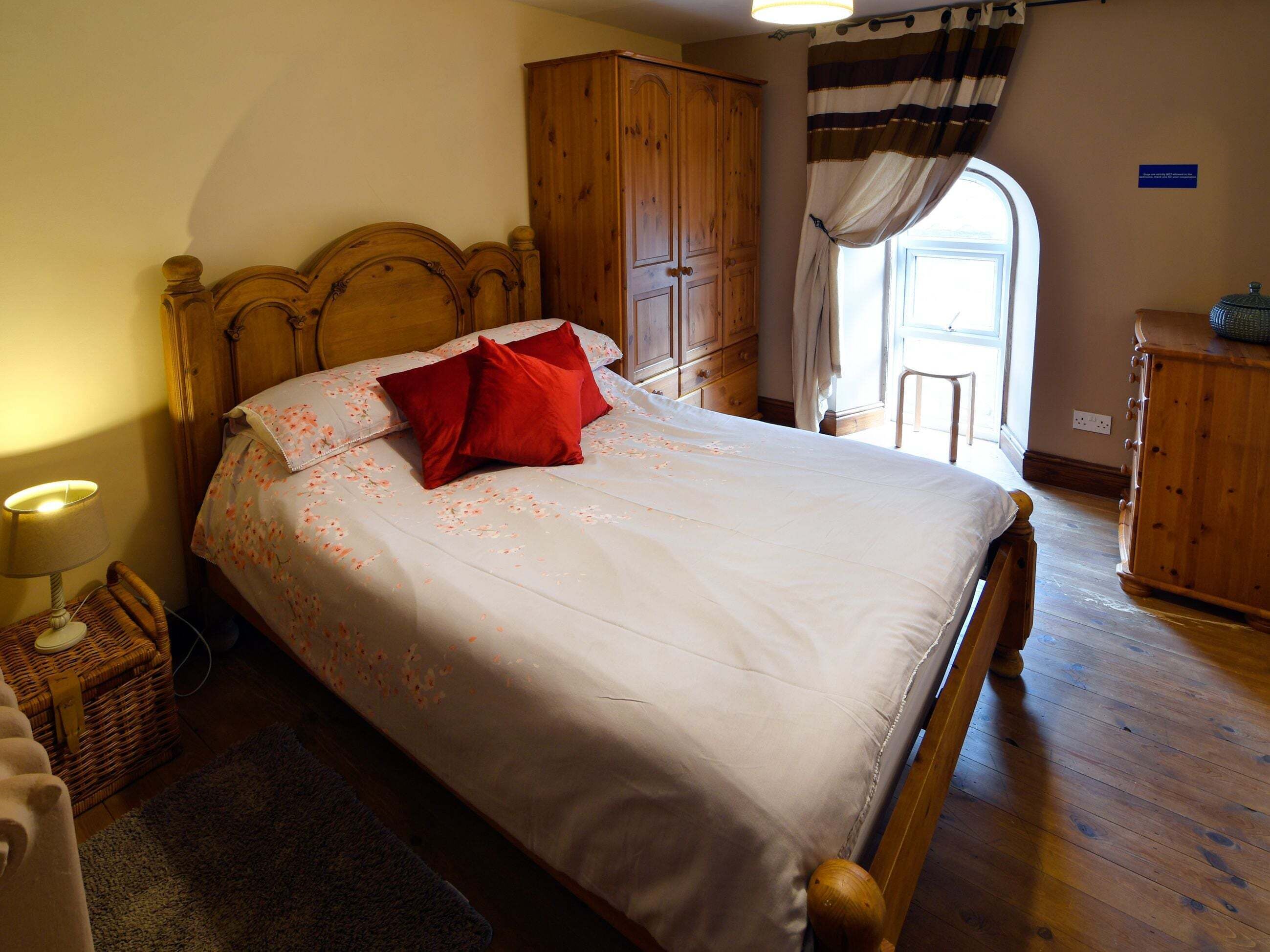 Een slaapkamer met een houten bed, nachtkastje, rieten stoel, houten kledingkast en een raam met gordijnen.