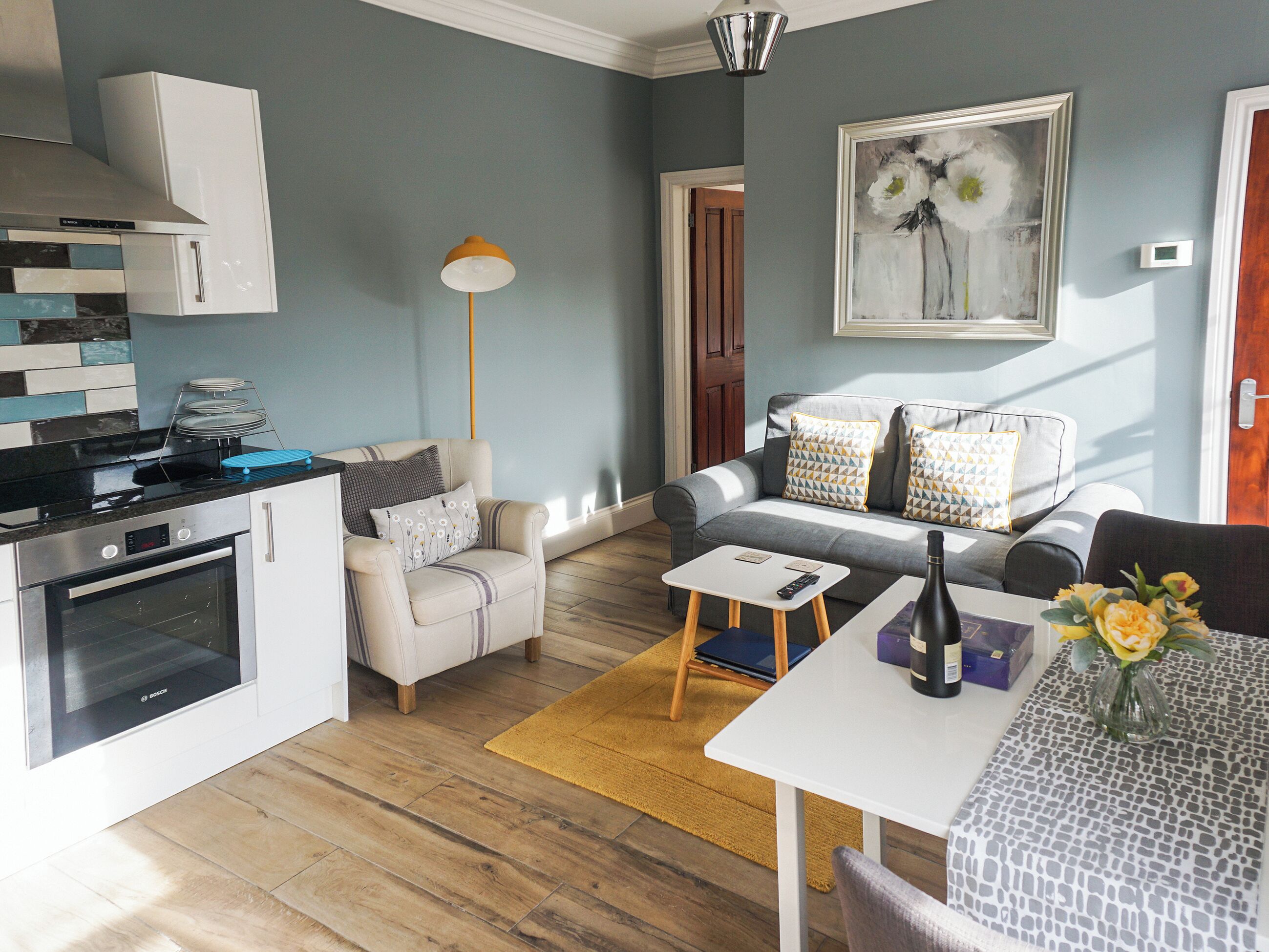 A modern kitchen with a Bosch oven, a grey sofa with patterned cushions, and a dining area with a white table and a vase of flowers.