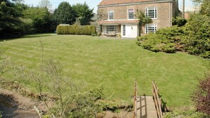 A large, well-maintained lawn with a wooden bridge leading to a brick house with multiple windows and a chimney. - Greenlands Farmhouse (Barmby Moor, York)