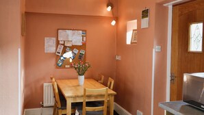 A dining area with a wooden table, chairs, a vase with flowers, a bulletin board, and a kitchen area with a countertop and appliances.