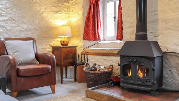 Un salón acogedor con un sillón de cuero, una mesa de madera con una lámpara, una cesta de leña y un estufa de leña.