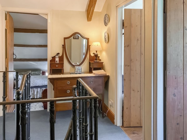 Une chambre avec une commode en bois, un grand miroir, une table de chevet avec une lampe et un escalier menant à l’étage supérieur.