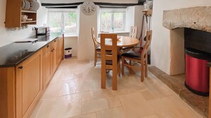 Une cuisine avec des meubles de rangement en bois, une table ronde et des chaises. Il y a une horloge au mur et une fenêtre donnant sur un paysage verdoyant.