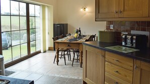 Une cuisine avec des meubles en bois, une table à manger pour quatre personnes, un réfrigérateur et une vue sur l’extérieur grâce à de grandes fenêtres.