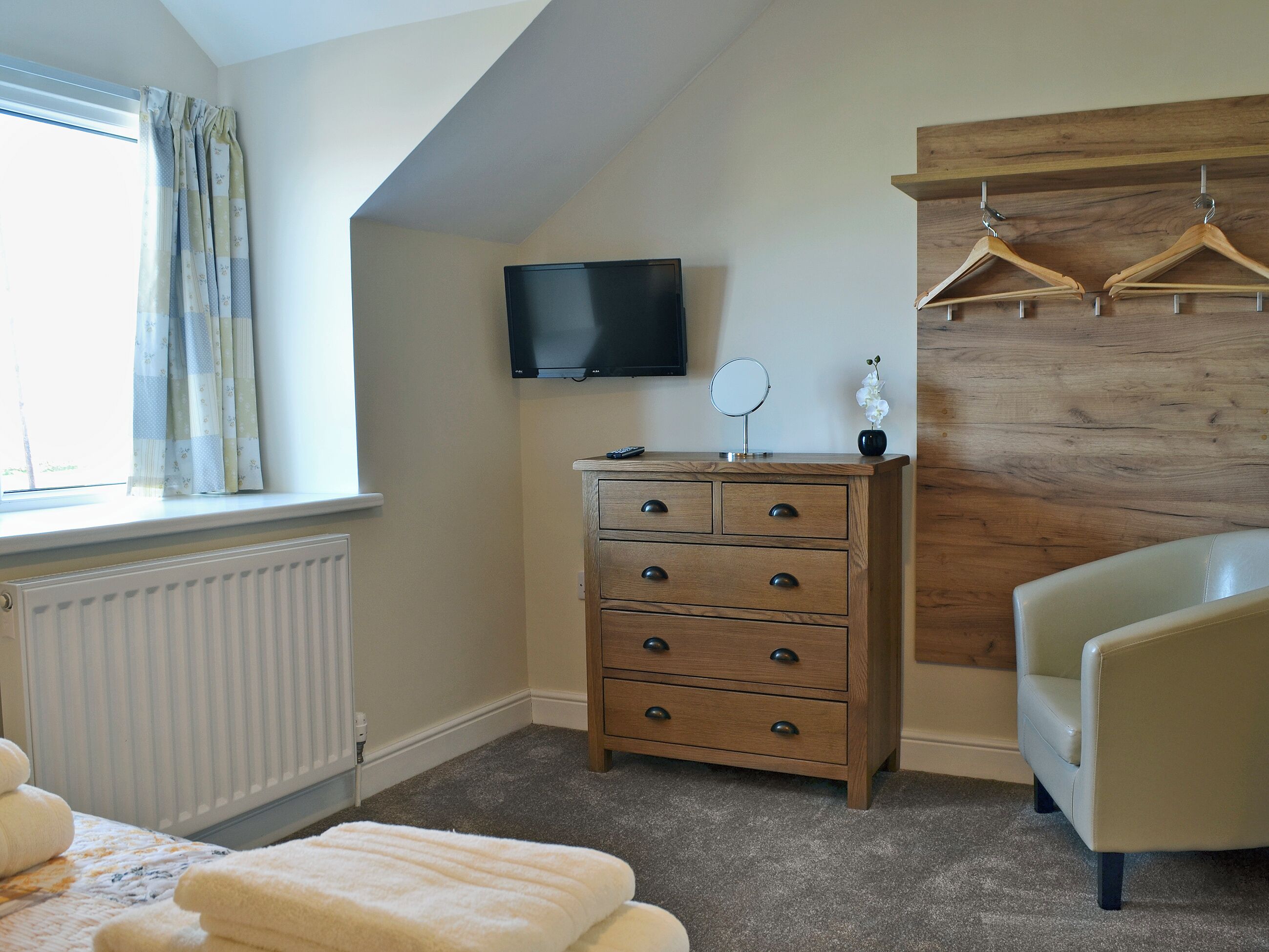 A bedroom with a wooden dresser, a television mounted on the wall, a mirror, and a chair.