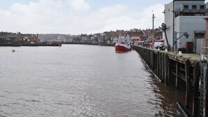 A waterfront view with a red boat docked at a pier, buildings along the shore, and a cloudy sky. - 2 bedroom accommodation in Whitby (Whitby)