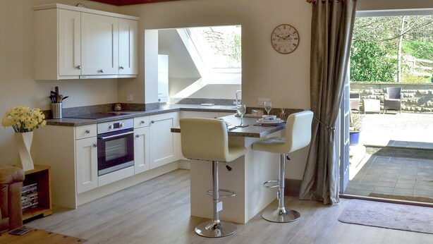 A modern kitchen with white cabinets, a built-in oven, and a dining area with two bar stools. - 1 bedroom accommodation in Baslow, near Bakewell (Baslow, near Bakewell)