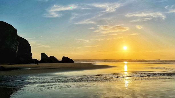 Una playa con un atardecer, cielo despejado y formaciones rocosas.