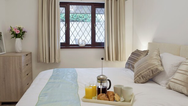 Een netjes opgemaakt bed met een ontbijttafel met croissants, koffie en sap. Een raam met gordijnen en uitzicht op groen is zichtbaar.