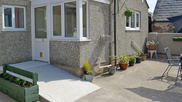Ein Haus mit kleiner Veranda, einem Fenster und einer Tür. Übertöpfe mit Pflanzen und eine Bank sind ebenfalls vorhanden.