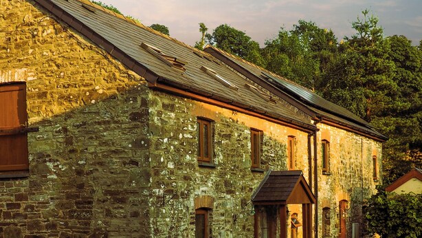 A rustic stone building with a sloped roof, wooden windows, and a small porch area with a chair and table. - 3 bedroom accommodation in Llanybri, near Carmarthen (Llanybri, near Carmarthen)
