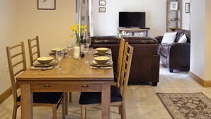 A dining area with a wooden table set for four, surrounded by chairs. A brown leather sofa and a television are visible in the background. - 2 bedroom accommodation in Spring Gardens, Frome (Spring Gardens, Frome)