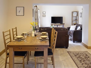 A dining area with a wooden table set for four, surrounded by chairs. A brown leather sofa and a television are visible in the background.