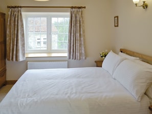 A bedroom with a large bed, a window with curtains, a bedside table with a vase of flowers, and a radiator.