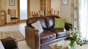 A living room with a brown leather sofa, two chairs, a coffee table with a book and flowers, a dining area with a table and chairs, and a kitchen area with cabinets and a fire extinguisher. - 2 bedroom accommodation in Spring Gardens, Frome (Spring Gardens, Frome)