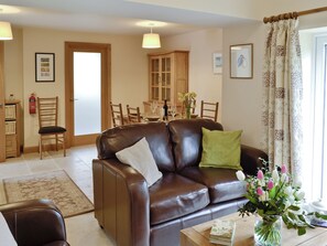 A living room with a brown leather sofa, two chairs, a coffee table with a book and flowers, a dining area with a table and chairs, and a kitchen area with cabinets and a fire extinguisher.
