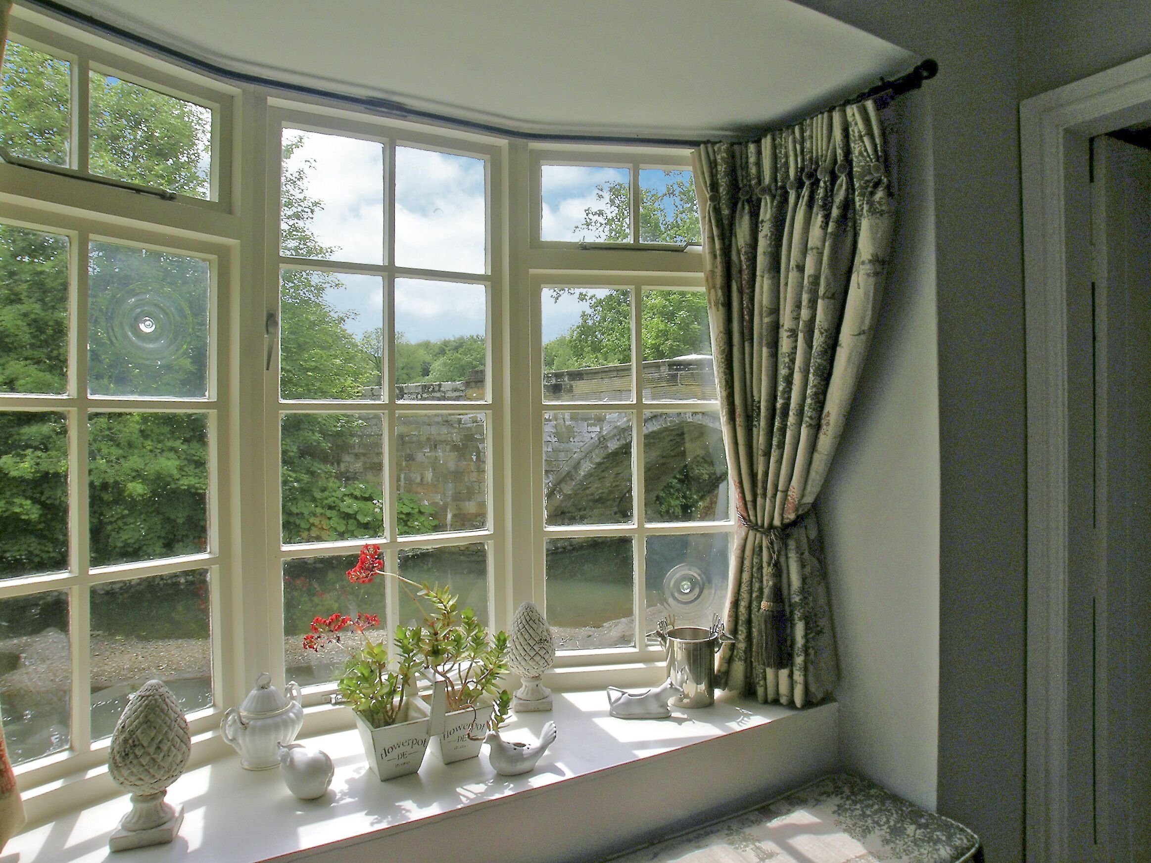 A bay window with multiple panes, a white table with decorative items, and a view of greenery and a stone structure outside.
