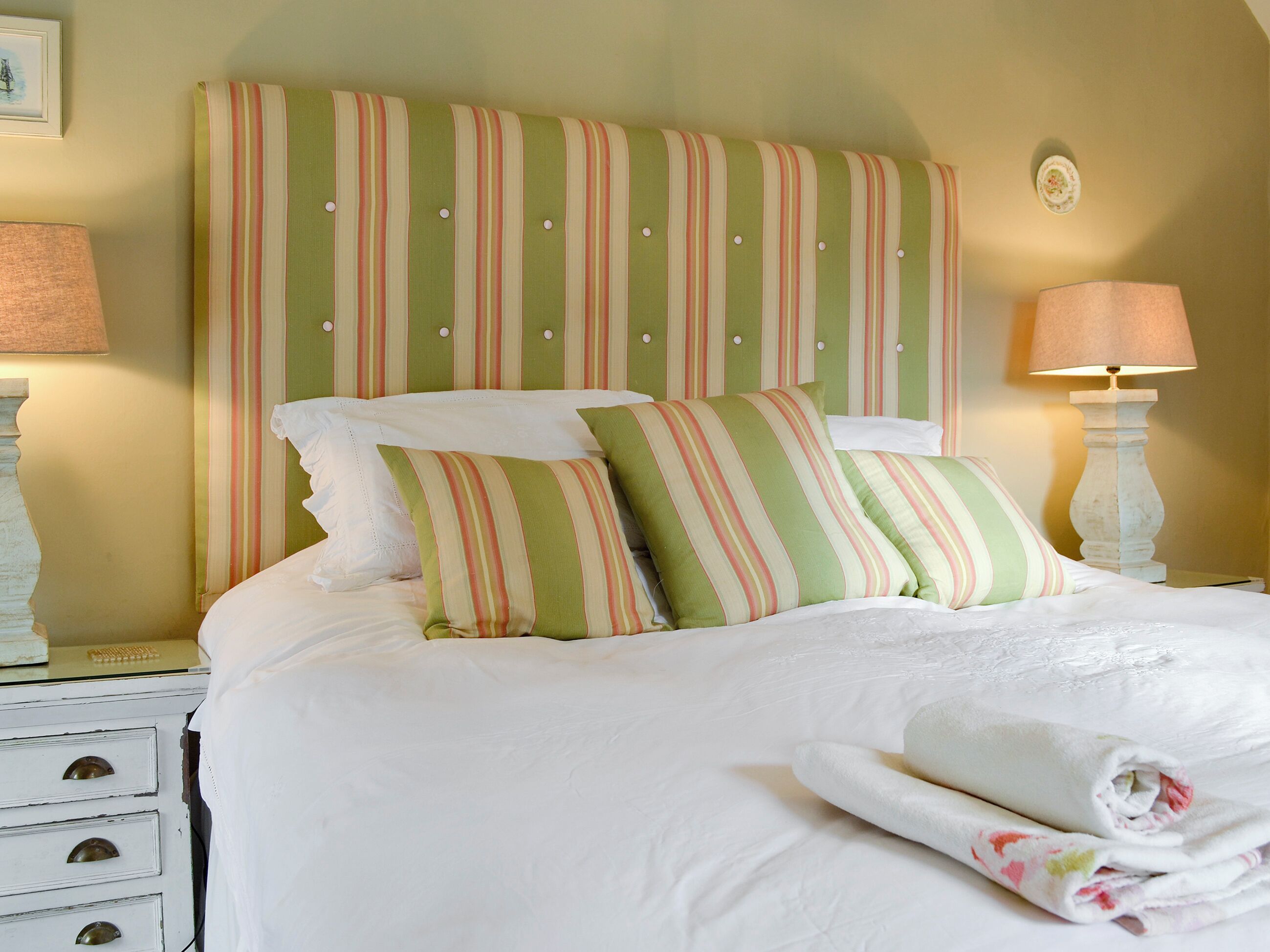 A neatly made bed with striped pillows and a headboard, a bedside table with a lamp, and a dresser with drawers.