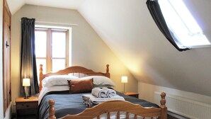 A bedroom with a sloped ceiling, a wooden bed, two bedside tables, a window with curtains, and a radiator. - Dusty's Stable - E4751 (Ebrington, near Chipping Campden)