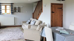 A cozy living room with a sofa, a dining table with a polka dot tablecloth, a wooden staircase, and a wooden door. - Dusty's Stable - E4751 (Ebrington, near Chipping Campden)