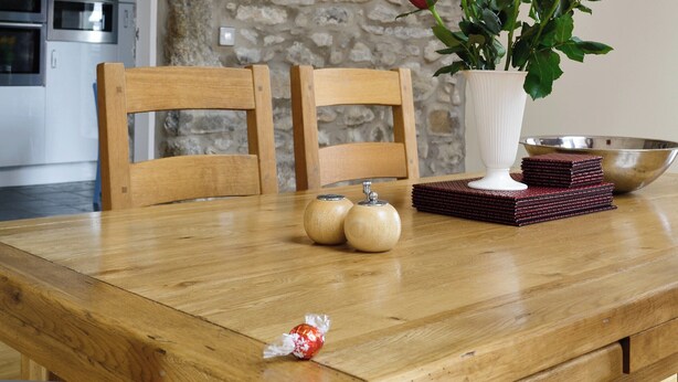 Una mesa de madera para comer con un jarrón de rosas rojas, dos frascos pequeños y una pila de posavasos.