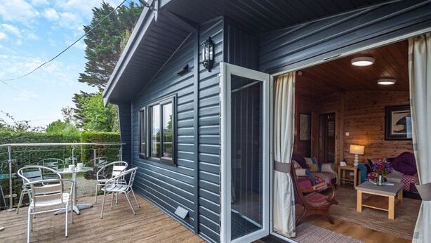 Une terrasse en bois avec des chaises et une table en métal, une porte-fenêtre coulissante donnant sur un intérieur chaleureux avec un canapé et une lampe.