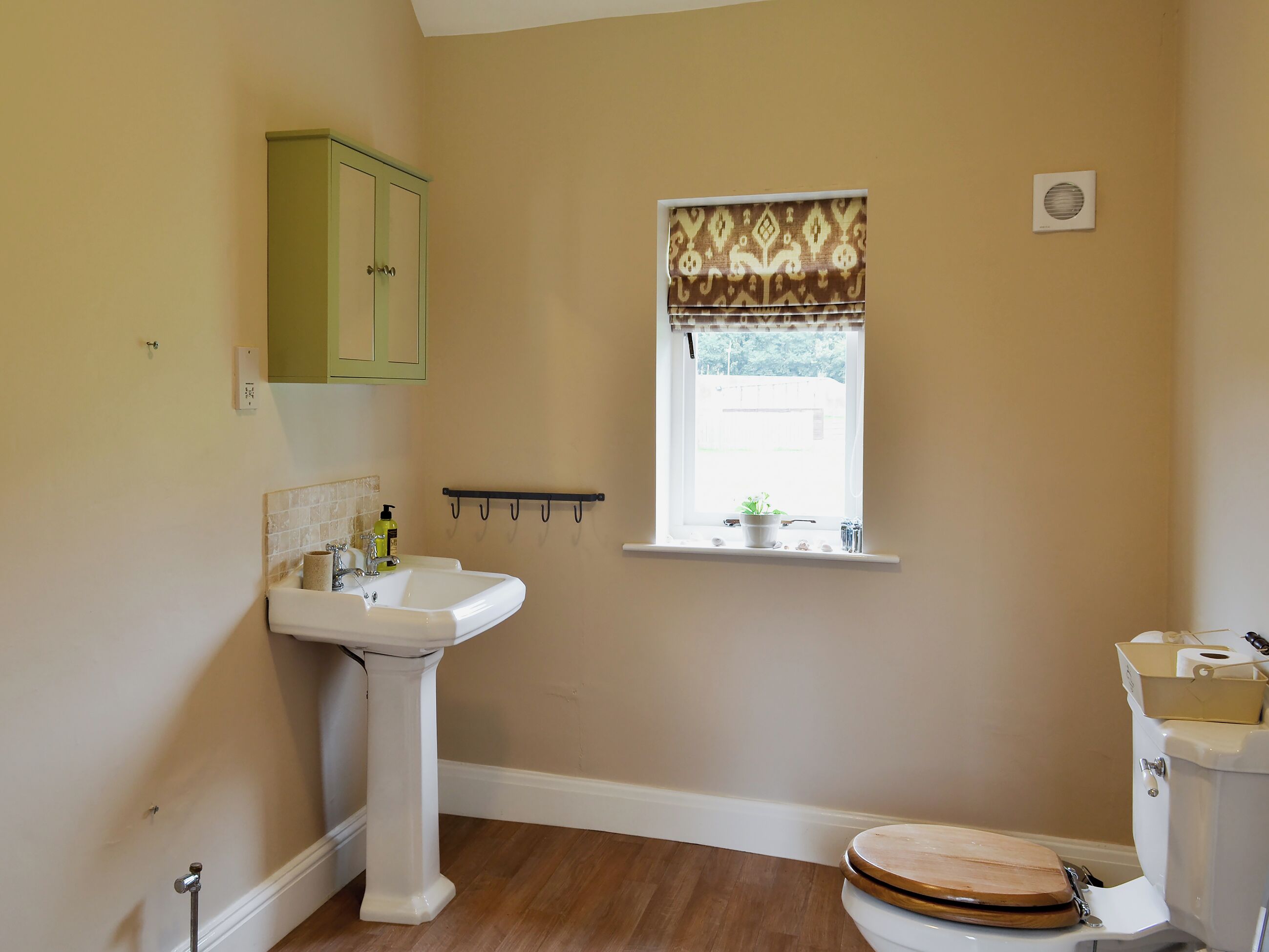 A bathroom with a pedestal sink, a toilet with a wooden seat, a window with a patterned blind, and a small shelf with a plant.