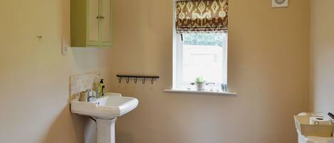 A bathroom with a pedestal sink, a toilet with a wooden seat, a window with a patterned blind, and a small shelf with a plant.
