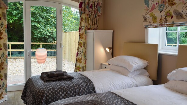 A bedroom with two beds, a wardrobe, a desk with a lamp, and a view of the garden through a glass door.