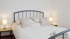 Een net opgemaakt bed met een zwart metalen hoofdbord, twee nachtkastjes met lampen, en een witte muur.