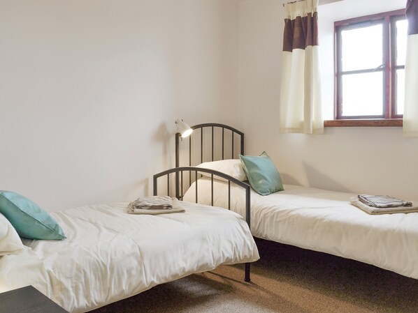 Une chambre avec deux lits simples, une table de chevet avec une lampe, une fenêtre avec des rideaux et un sol recouvert d’une moquette.