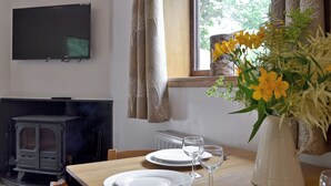 Een eethoek met een tafel voor twee personen, een aan de muur bevestigde televisie, een houtkachel en een raam met gordijnen.