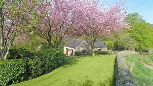 Een stenen huis met rieten dak, omgeven door een verzorgd gazon en bloeiende bomen.