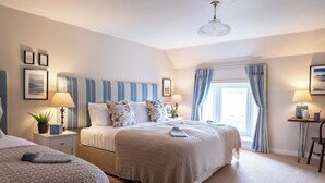 Ein Schlafzimmer mit zwei Betten, einem großen Fenster mit Vorhängen, einem Nachttisch mit Lampe und gerahmten Bildern an der Wand.