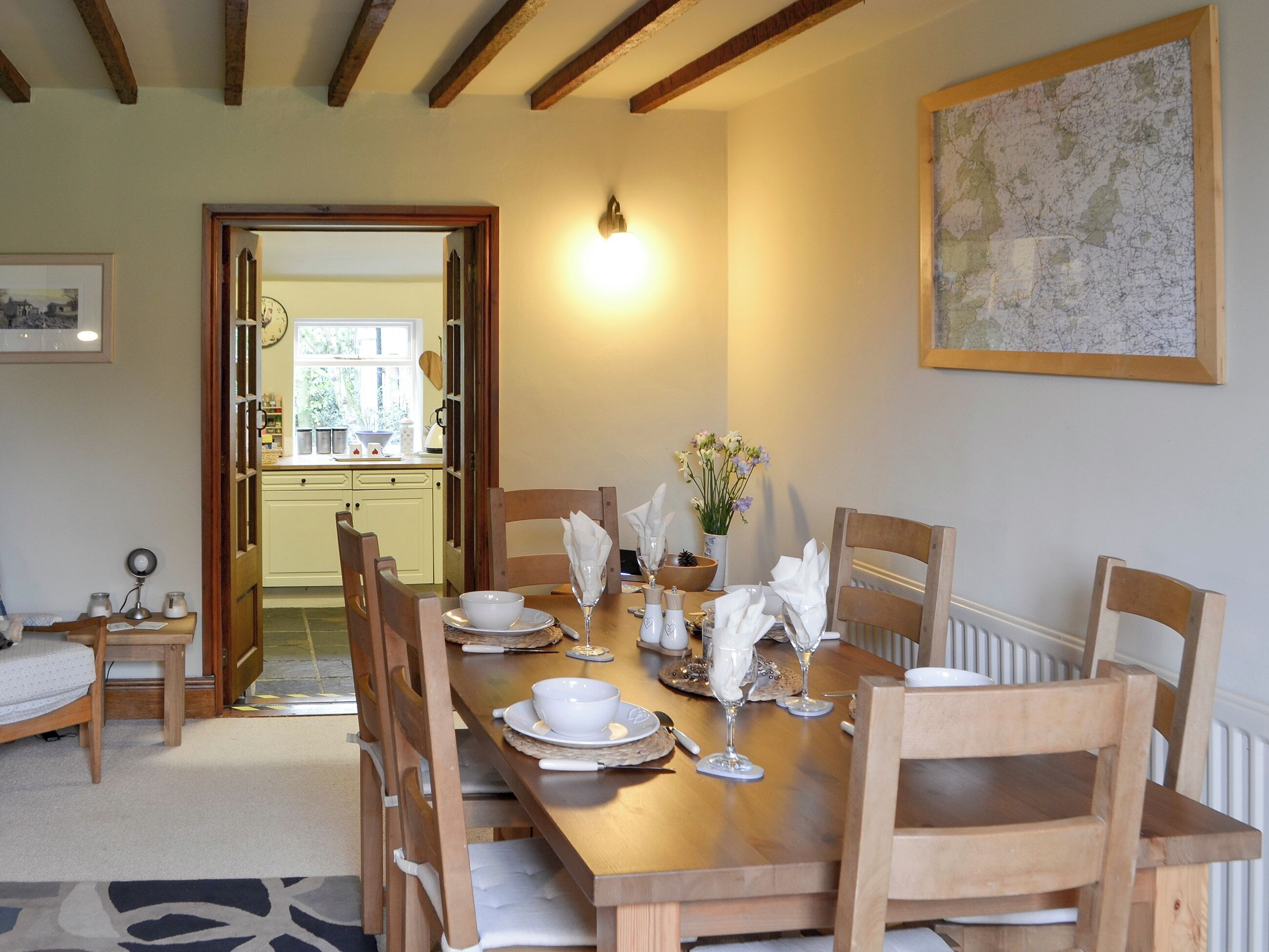 A dining room with a wooden table set for four, surrounded by wooden chairs. There is a small table with a chair to the left, a framed picture on the wall, and a map on the right.