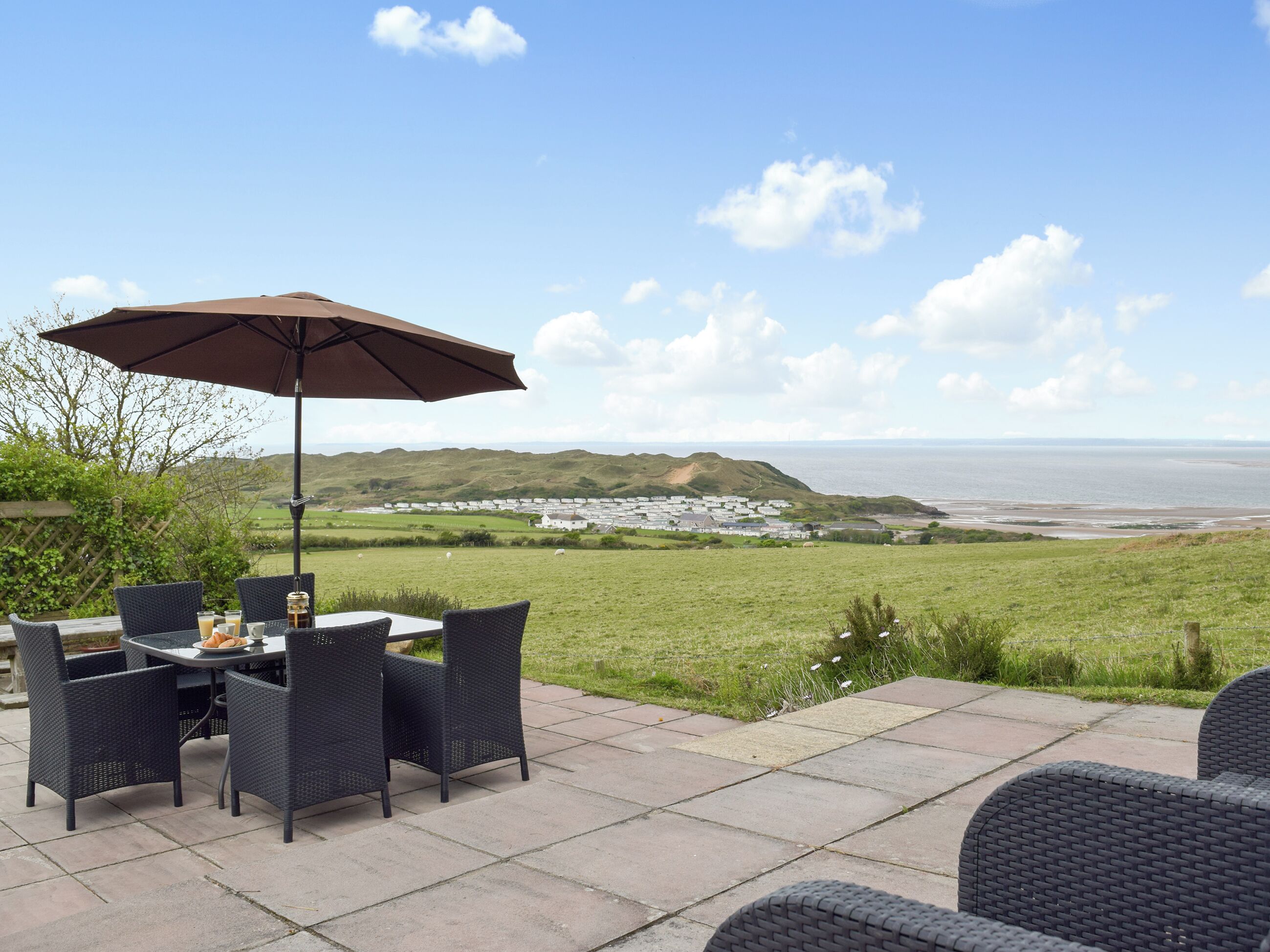 An outdoor patio with a table and chairs, a large umbrella, and a view of the sea and distant buildings.