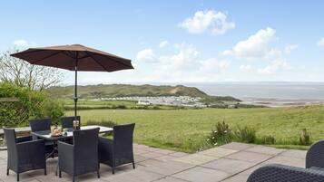 An outdoor patio with a table and chairs, a large umbrella, and a view of the sea and distant buildings.